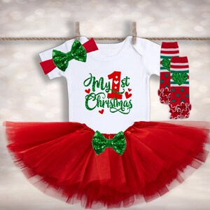 May include: A red tulle tutu skirt with a white onesie that says "My 1st Christmas" in green glitter lettering. The onesie has a red number one with a green heart on it. The outfit is completed with a green glitter bow and red and green leg warmers with white stripes and green hearts.