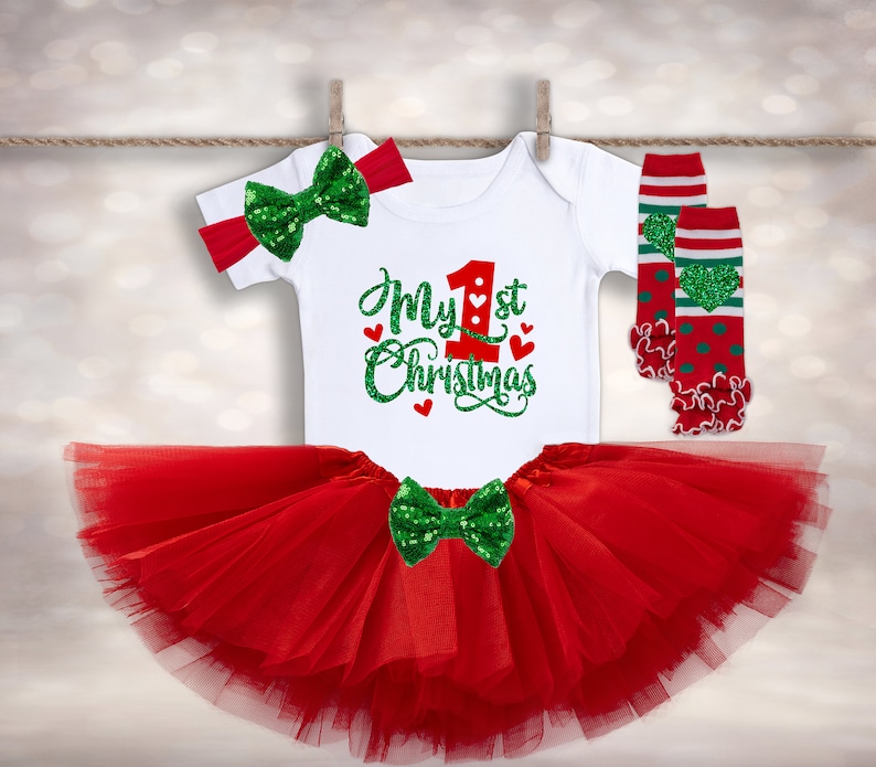 May include: A red tulle tutu skirt with a white onesie that says "My 1st Christmas" in green glitter lettering. The onesie has a red number one with a green heart on it. The outfit is completed with a green glitter bow and red and green leg warmers with white stripes and green hearts.