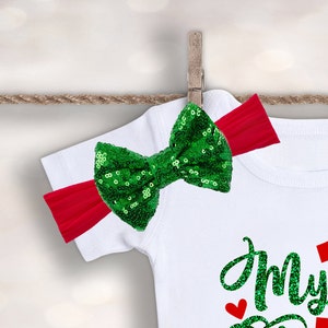 May include: A white onesie with the text "My First Christmas" in green glitter. A red headband with a green sequin bow is attached to the onesie.