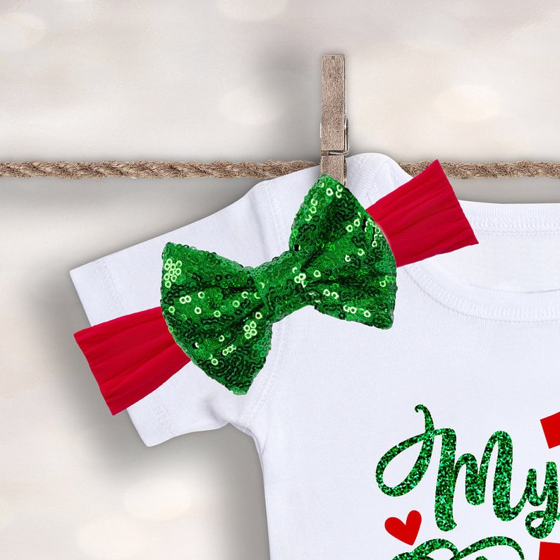 May include: A white onesie with the text "My First Christmas" in green glitter. A red headband with a green sequin bow is attached to the onesie.