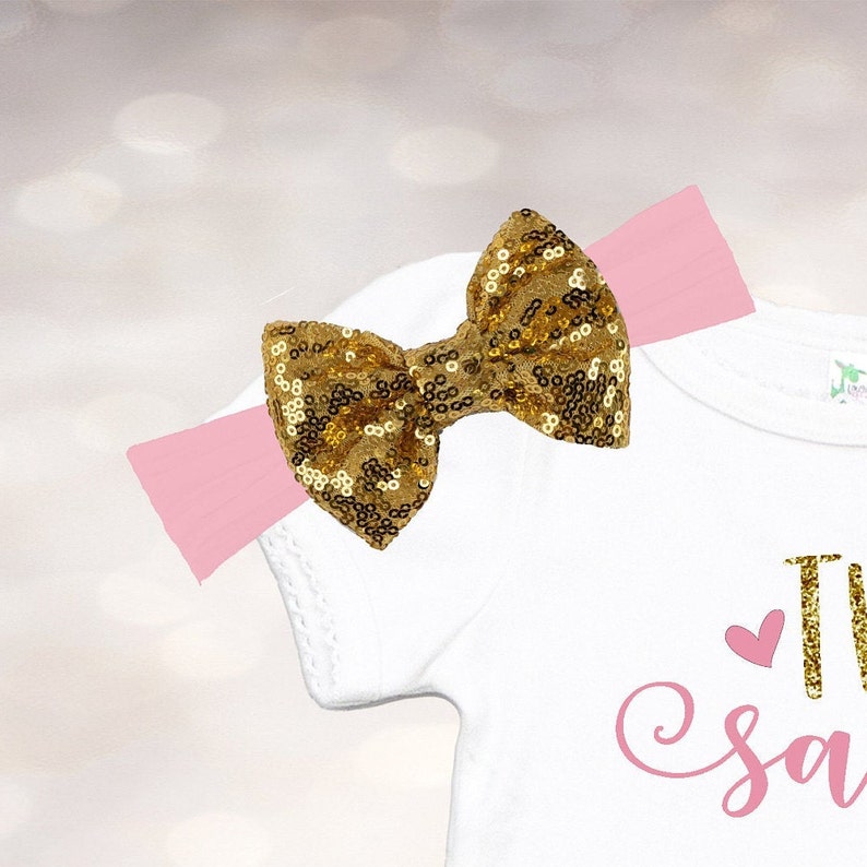 May include: A white onesie with pink text that says "Two Sweet" and a pink heart. A gold sequin bow is attached to a pink headband.