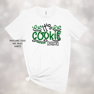 May include: White t-shirt with green glitter text that reads "It's Cookie Season".