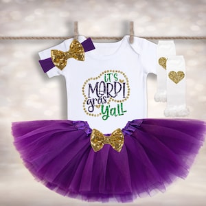 May include: Purple tulle tutu with a gold bow, a white onesie with the text "It's Mardi Gras Y'all" in gold glitter, and a white headband with a gold bow.