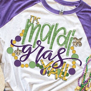 May include: A white and purple t-shirt with the text "It's Mardi Gras Y'all" in green, purple, and gold. The text is surrounded by purple, green, and gold circles and fleur-de-lis.