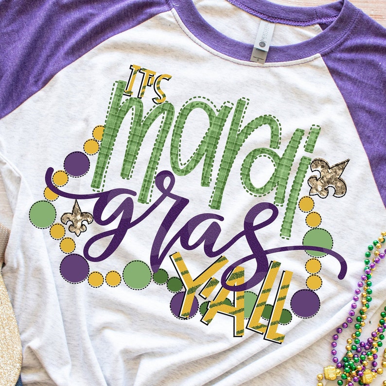 May include: A white and purple t-shirt with the text "It's Mardi Gras Y'all" in green, purple, and gold. The text is surrounded by purple, green, and gold circles and fleur-de-lis.