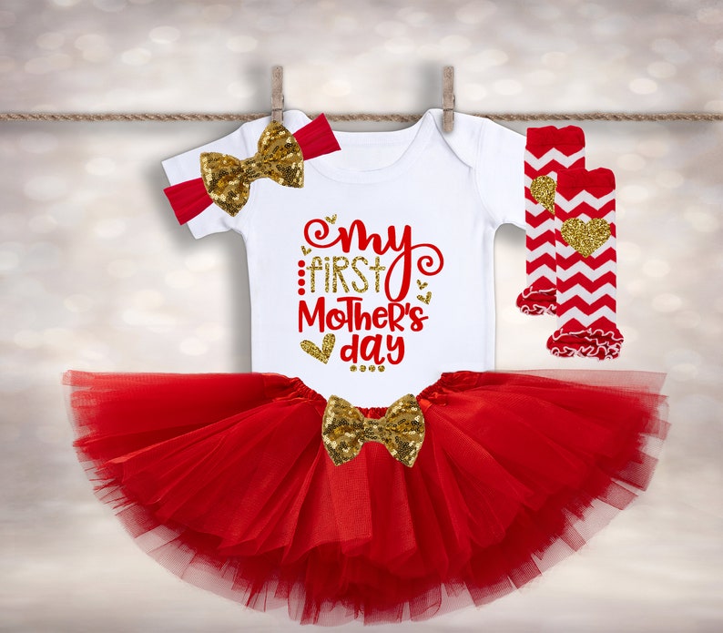 Puede incluir: Conjunto de beb&eacute; con un body blanco con texto rojo y dorado "My First Mother's Day". El conjunto incluye un tut&uacute; rojo, una diadema y calcetines. Decorado con lazos dorados con lentejuelas y calcetines con estampado de chevron rojo y blanco.