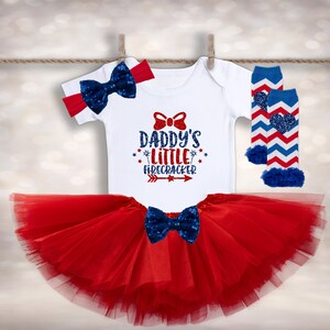 May include: Red, white, and blue baby outfit with a white bodysuit that says "Daddy's Little Firecracker" with a red bow, a red tulle tutu, and blue and white striped leg warmers with a red heart.