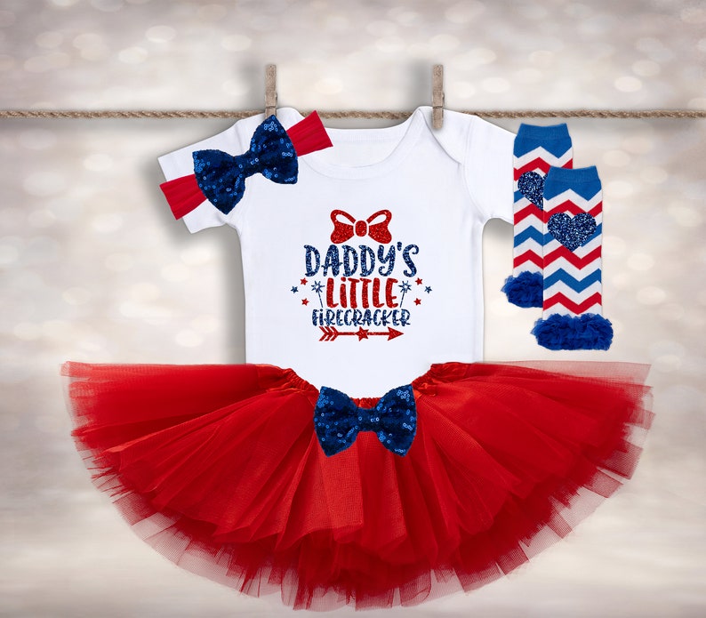 May include: Red, white, and blue baby outfit with a white bodysuit that says "Daddy's Little Firecracker" with a red bow, a red tulle tutu, and blue and white striped leg warmers with a red heart.