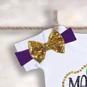 May include: A white baby onesie with a gold glitter 'Mardi Gras' design and a purple headband with a gold sequin bow.