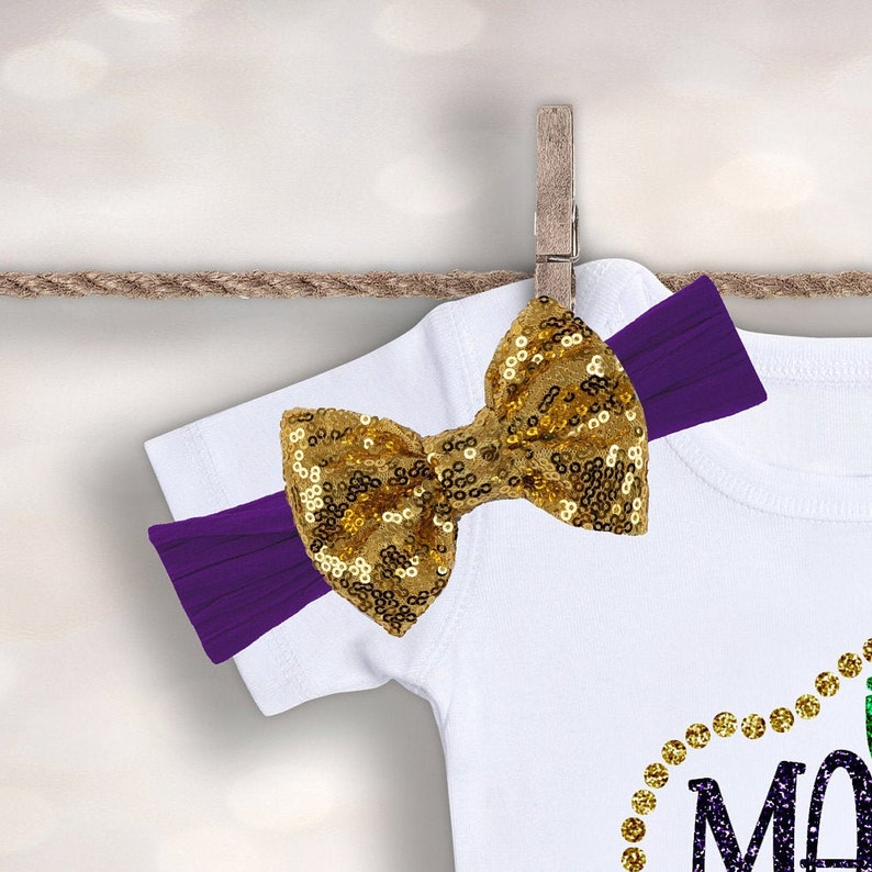 May include: A white baby onesie with a gold glitter 'Mardi Gras' design and a purple headband with a gold sequin bow.