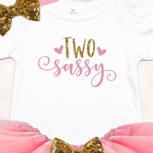 May include: A white onesie with pink glitter text that says "TWO sassy" with two pink hearts. The onesie is paired with a pink tulle skirt and a gold glitter bow.