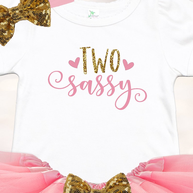 May include: A white onesie with pink glitter text that says "TWO sassy" with two pink hearts. The onesie is paired with a pink tulle skirt and a gold glitter bow.