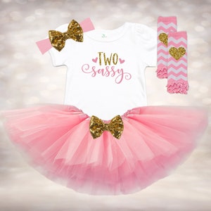 May include: Pink tutu with a gold glitter bow, a white onesie with the text "TWO sassy" in pink, and pink and white striped leg warmers with a gold glitter heart.