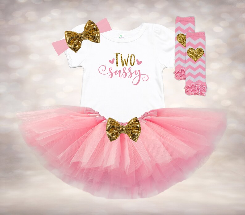 May include: Pink tutu with a gold glitter bow, a white onesie with the text "TWO sassy" in pink, and pink and white striped leg warmers with a gold glitter heart.