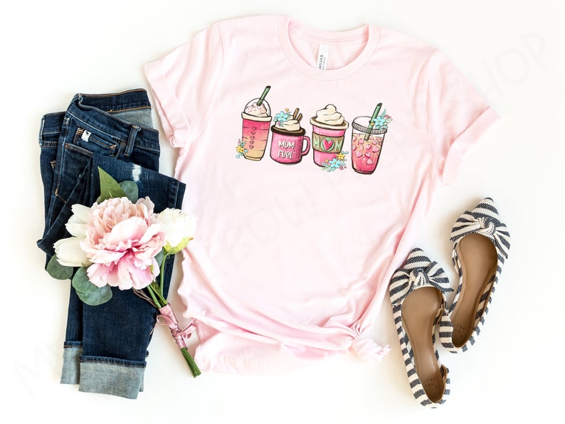 Op de afbeelding: Roze T-shirt met een afbeelding van vier verschillende koffiedrankjes. De drankjes zijn versierd met bloemen en de tekst "Mom Fuel". Het shirt wordt gecombineerd met een blauwe jeans, een boeket roze bloemen en gestreepte ballerina's.