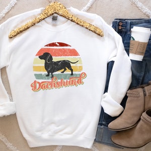 May include: White sweatshirt with a vintage-style graphic of a dachshund silhouette in front of a sunset. The text "Dachshund" is written in a retro font below the image.