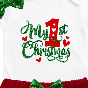 May include: White onesie with green glitter text that says "My 1st Christmas". The onesie has a red number one with white hearts. The onesie is paired with a red skirt and a green sequin bow.