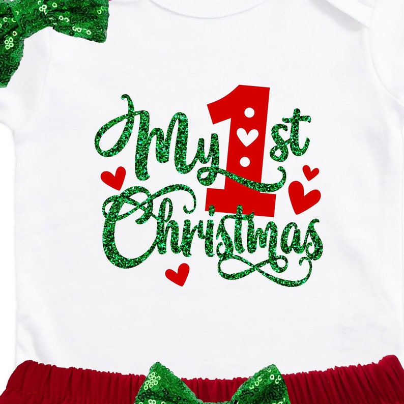 May include: White onesie with green glitter text that says "My 1st Christmas". The onesie has a red number one with white hearts. The onesie is paired with a red skirt and a green sequin bow.