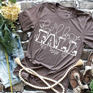 May include: A brown t-shirt with a white graphic that says "hello fall" with fall in a larger font and surrounded by small white floral designs.