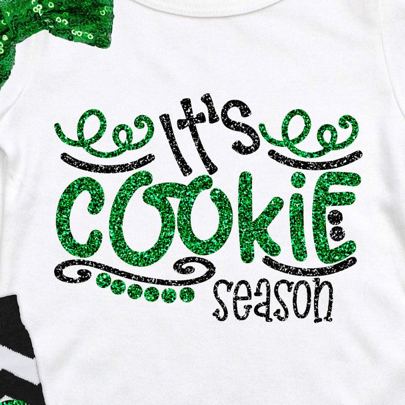 May include: White shirt with green glitter text that reads "It's Cookie Season".