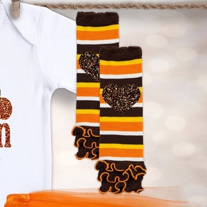 May include: A pair of brown, orange and white striped leg warmers with a brown heart-shaped glitter design. The leg warmers have a ruffled edge.