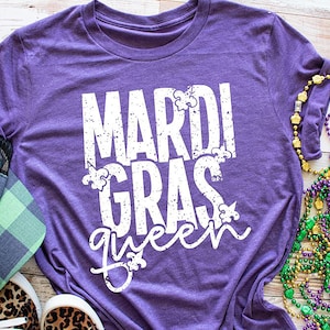 May include: Purple t-shirt with the text "Mardi Gras queen" in white with a distressed look. The shirt is surrounded by Mardi Gras beads and a green and black plaid hat.