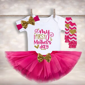 Puede incluir: Un conjunto de ropa de beb&eacute; que incluye un body blanco con "My First Mother's Day" en rosa y dorado, un tut&uacute; rosa, una diadema con lazo de lentejuelas doradas, un lazo de lentejuelas doradas y calentadores de piernas rosas y blancos. El conjunto est&aacute; colgado.