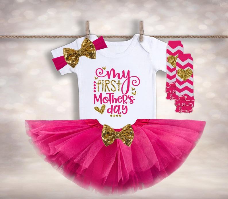 Puede incluir: Un conjunto de ropa de beb&eacute; que incluye un body blanco con "My First Mother's Day" en rosa y dorado, un tut&uacute; rosa, una diadema con lazo de lentejuelas doradas, un lazo de lentejuelas doradas y calentadores de piernas rosas y blancos. El conjunto est&aacute; colgado.