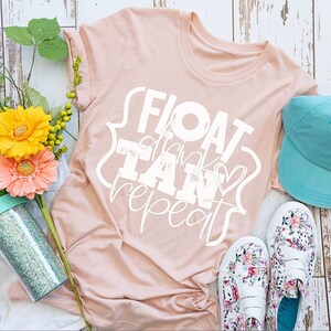 以下が含まれることがあります： 白い文字で「FLOAT drink TAN repeat」と書かれたピンクのTシャツ。白い木製の表面に、ターコイズブルーの野球帽、花柄の靴、透明なキラキラのタンブラーと一緒に置かれています。