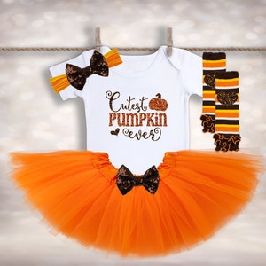 May include: Orange tulle tutu skirt, white bodysuit with "Cutest Pumpkin Ever" text and a pumpkin graphic, and orange and brown striped leg warmers with a brown bow headband.