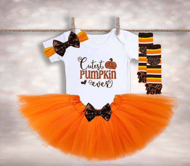 May include: Orange tulle tutu skirt, white bodysuit with "Cutest Pumpkin Ever" text and a pumpkin graphic, and orange and brown striped leg warmers with a brown bow headband.