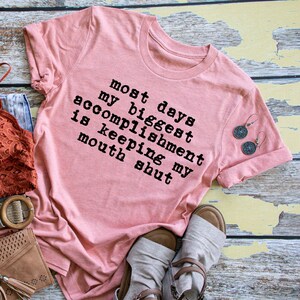 May include: A pink t-shirt with the text "most days my biggest accomplishment is keeping my mouth shut" printed in black.