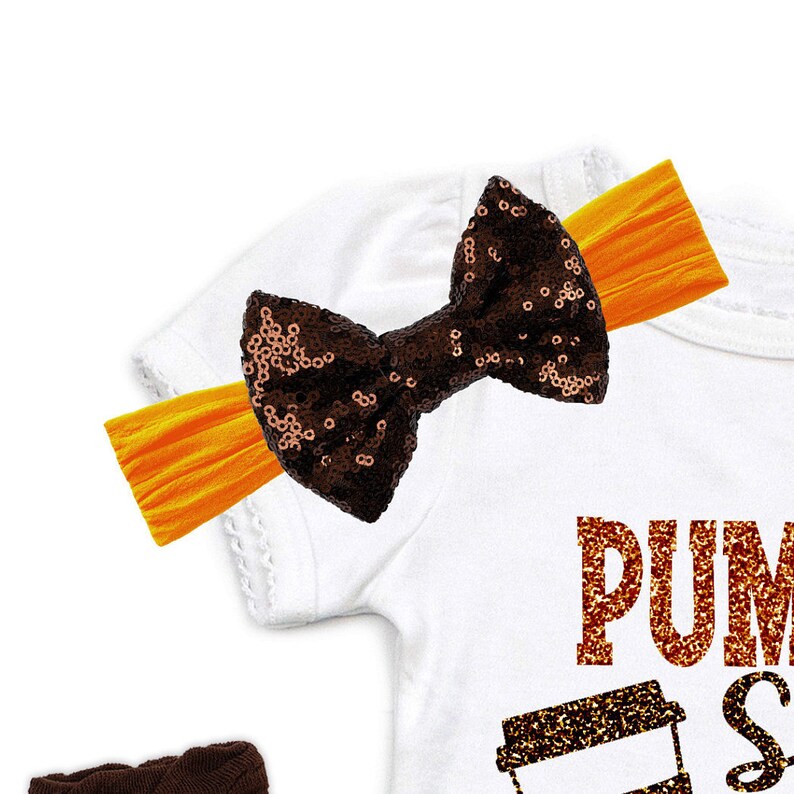 May include: A white baby bodysuit with the words "PUMPKIN SPICE" in orange glitter lettering. A brown sequined bow is attached to an orange headband. The bodysuit has short sleeves and a coffee cup graphic.