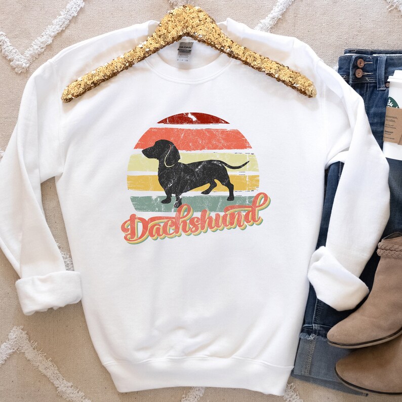 May include: White sweatshirt with a vintage-style graphic of a dachshund silhouette in front of a sunset. The text "Dachshund" is written in a retro font below the image.