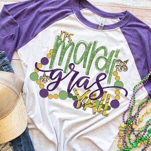 May include: A white and purple raglan t-shirt with the text "It's Mardi Gras Y'all" in green and gold lettering. The shirt is decorated with green, gold, and purple beads and a fleur-de-lis.
