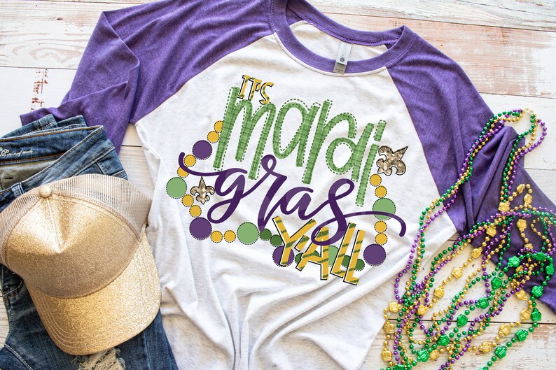 May include: A white and purple raglan t-shirt with the text "It's Mardi Gras Y'all" in green and gold lettering. The shirt is decorated with green, gold, and purple beads and a fleur-de-lis.