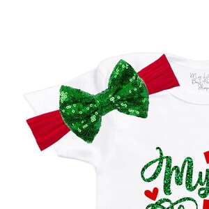 May include: White baby onesie with green glitter text that says "My First Christmas". The onesie has a green sequin bow on the shoulder with a red headband.
