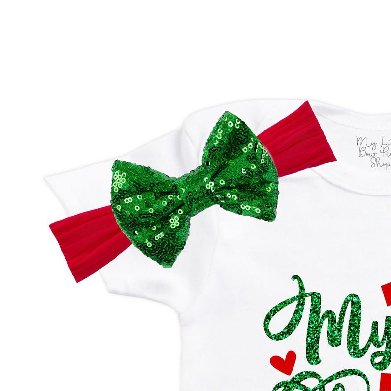 May include: White baby onesie with green glitter text that says "My First Christmas". The onesie has a green sequin bow on the shoulder with a red headband.