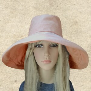 Puede incluir: Un sombrero de sol de ala ancha de color rosa melocot&oacute;n con un tono ligeramente m&aacute;s oscuro alrededor del borde. El sombrero est&aacute; hecho de una tela suave y ligera y tiene un dise&ntilde;o simple y elegante.