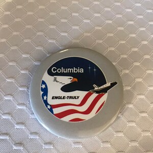 May include: A round, silver button with a white and red American flag, a white space shuttle, and a bald eagle. The text "Columbia" and "ENGLE-TRULY" are printed on the button.