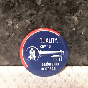 May include: A blue and red round button with a white key and a space shuttle. The text on the button reads "QUALITY... key to leadership in space".