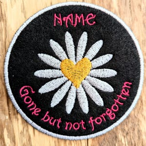 May include: A black embroidered patch with a white daisy and a yellow heart in the centre. The text "NAME" is at the top and "Gone but not forgotten" is around the bottom of the patch.