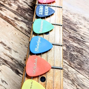 May include: A close-up of six guitar picks on a guitar neck. The picks are different colors and have different names written on them: "Gourler", "Ziggy", "Amarante", "Motorhead", "Milkshake", and "Malina".