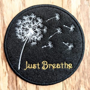 May include: A black round felt patch with a white embroidered dandelion and the words "Just Breathe" in gold embroidery.