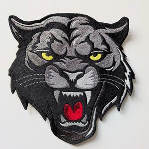 May include: Embroidered patch of a snarling panther head. The patch is black and grey with yellow eyes and a red mouth. The design is detailed with visible stitching.