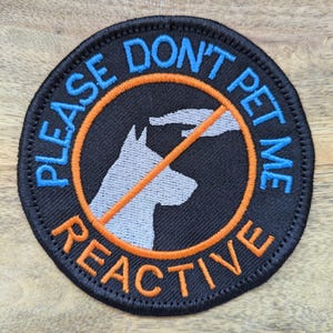 Peut inclure: Écusson rond noir avec texte bleu et orange et silhouette de chien blanche. Le texte indique « PLEASE DON'T PET ME REACTIVE » avec une main et un chien barrés. L'écusson est conçu pour être cousu sur des vêtements ou accessoires.