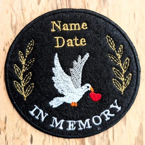 Peut inclure: Patch commémorative en feutre noir avec une colombe blanche portant un cœur rouge. Le patch est orné de feuilles brodées dorées de chaque côté et du texte "Nom Date En Mémoire".