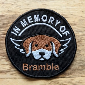 May include: A black embroidered patch with a white border and the text "In Memory Of" in a white arc above a brown and white cartoon dog with wings. The text "Bramble" is embroidered in brown below the dog.