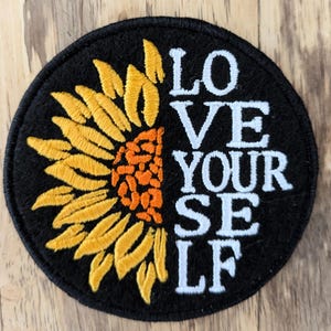 May include: A black and yellow embroidered patch featuring a sunflower design and the text "LOVE YOURSELF" in white.