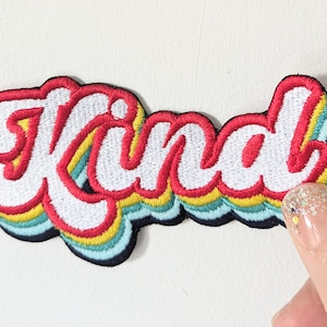 May include: A colourful embroidered patch with the word "Kind" in a retro font. The patch has a rainbow design with red, yellow, blue, and green colours.